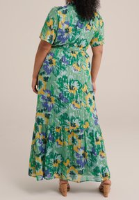 Maxiklänning i grönt tyg med blommönster i gult och lila. Har korta ärmar, knytning i midjan och en flerlagerskjol.