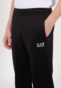 Zwarte joggingbroek met een elastische tailleband en zijzakken. Bevat een witte geborduurde logo op de dij. Zacht stofgevoel.