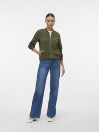 Veste bomber vert olive avec un col rond et une fermeture éclair devant, associée à un jean bleu évasé et des chaussures blanches. Texture lisse, poches pratiques.