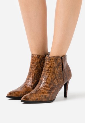 Bruine enkellaarsjes met een slangenhuidtextuur, spitse neus en smalle hoge hakken, gedragen op blote benen tegen een witte achtergrond.
