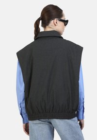 Gilet oversized grigio scuro con collo e orlo elastico, indossato sopra una camicia a maniche lunghe blu chiaro, caratterizzato da una texture di tessuto liscia.