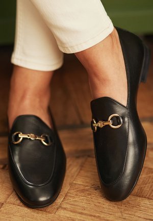 Svarta läderloafers med en slät textur, som har en guldfärgad hårdvaruaccent översta delen och en rund tåsdesign.