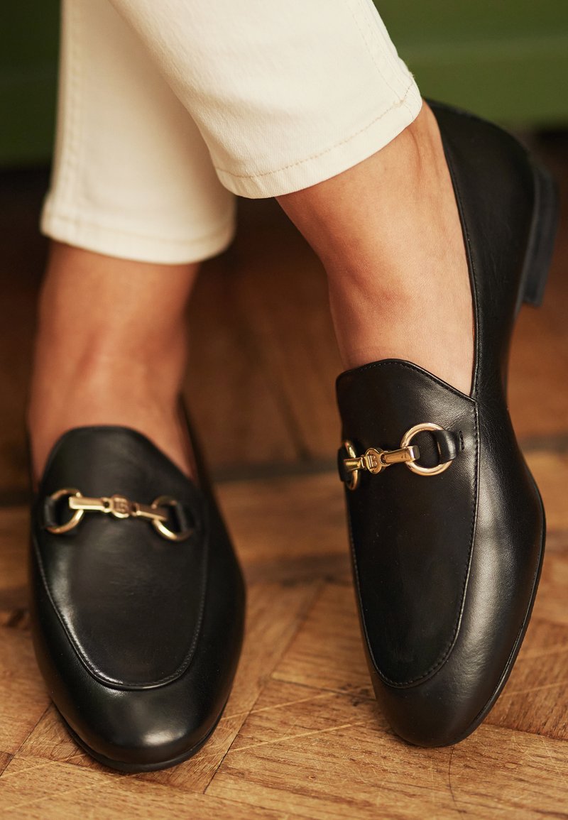 Sorte læder loafers med en glat tekstur, der har en guldfarvet hardwaredetalje over toppen og et rundt tådesign.