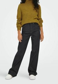 Dunkelblaue weit geschnittene Jeans, kombiniert mit einem senfgelben Rollkragenpullover mit gerippter Textur und langen Ärmeln, vor einem schlichten Hintergrund.