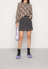 Femme portant un chemisier fleuri, une jupe en denim noire et des bottines noires avec des semelles épaisses lavande, tenant un sac à main lavande à côté d'elle.