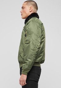 Alyvuogių žalios spalvos bombonešio striukė su juoda dirbtinio kailio apykakle, užtrauktuko kišene ant rankovės, apsiūtais rankogaliais ir apačia, bei lygiu satino audinio paviršiumi.