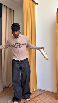 Νέος άνδρας με μαύρο σκουφάκι και ριγέ πουλόβερ με φερμουάρ, κρατώντας ένα λευκό αθλητικό παπούτσι στο ένα χέρι, στέκεται σε ξύλινο πάτωμα κοντά σε κίτρινη κουρτίνα.