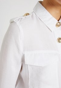 Camisa blanca, ligera, con cuello, dos bolsillos en el pecho y botones decorativos dorados en el hombro y el delantero. Textura suave.
