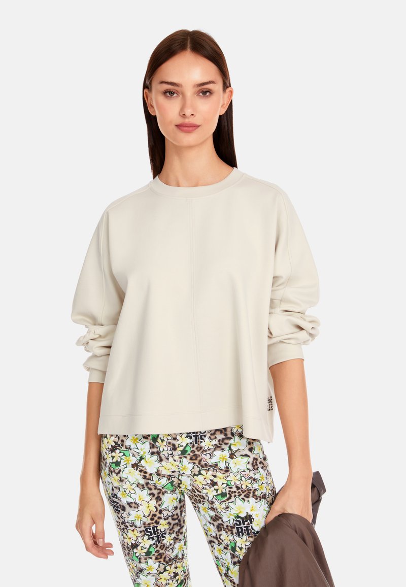 Hellgrauer, oversized Sweatshirt mit langen, gerafften Ärmeln und gestickten Details. Kombiniert mit gemusterten, bunten Leggings.