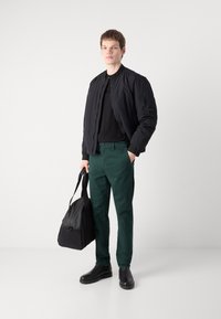 Cazadora bomber negra, camiseta negra, pantalones verdes y botas negras. Lleva una bolsa negra con cremallera. Diseño minimalista y colores sólidos.