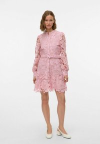 Robe en dentelle rose avec des manches longues, motif floral, devant à boutons, taille marquée par une ceinture et ourlet superposé en forme de vagues. Fini avec un tissu texturé.