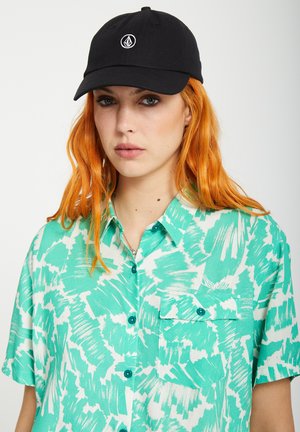 Femme aux longs cheveux orange portant une casquette noire et une chemise à motifs bleu sarcelle et blanc avec une poche poitrine.