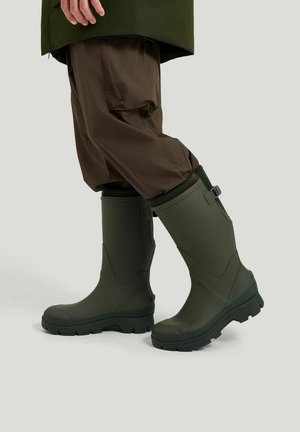 Person iført mørkegrønne gummistøvler og brune cargobukser, stående med den ene fod let løftet på en lys baggrund.
