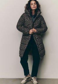 Pufová bunda s leopardím vzorom a vysokým golierom, so zapínaním na zips, textúrovanou látkou a bočnými vreckami, skombinovaná s tmavými džínsami a teniskami.