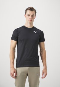 Černé sportovní tričko s krátkým rukávem s texturovaným povrchem a bílým logem Puma na hrudi, doplněné světlými khaki kalhotami.