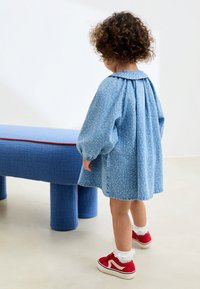 Lyseblå blomstrede denimkjole med puffede ærmer og Peter Pan-krave, kombineret med røde sneakers og hvide flæsebesatte sokker, set bagfra.