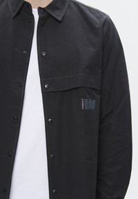 Black shirt jacket in a lightweight fabric, featuring snap buttons, a chest pocket, and a subtle logo detail near the pocket.
