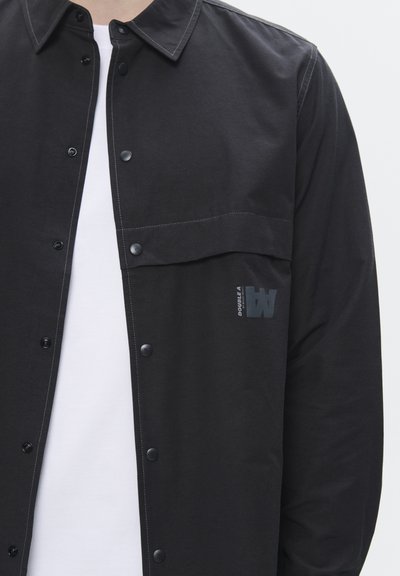 Schwarze Hemdjacke aus leichtem Material, mit Druckknöpfen, einer Brusttasche und einem dezenten Logodesign nahe der Tasche.