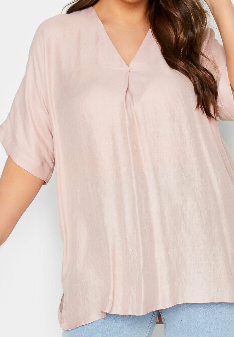 Blouse ample rose pâle à col en V avec manches courtes portée avec un jean bleu clair, montrée du milieu du torse au cou avec le bras droit levé.