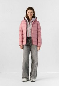 Veste rembourrée rose clair avec capuche, design matelassé et fermeture éclair, superposée sur une veste grise, associée à un jean large gris.