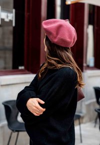 Tűnő rózsaszín beret, lekerekített formával és egy kis hurokkal a tetején, egy fekete pulcsit viselő személyen, akinek hullámos barna haja részben látható.