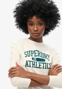 Jasno beżowa, długosleeve koszula z nadrukiem "SUPERDRY ATHLETICS 72" w ciemnej zieleni, okrągły dekolt, miękka tekstura, model z założonymi rękami.