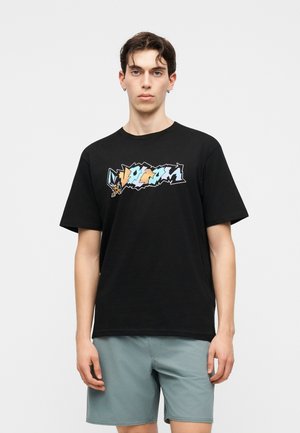 Νέος άνδρας με μαύρο T-shirt με πολύχρωμο αφηρημένο σχέδιο και ανοιχτό μπλε σορτς, στέκεται μπροστά σε απλό ανοιχτό φόντο.