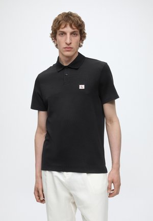 Νέος άνδρας με σγουρά μαλλιά φοράει μαύρο κοντομάνικο πόλο πουκάμισο με μικρό κόκκινο και λευκό λογότυπο στο στήθος, συνδυασμένο με λευκή παντελόνα.