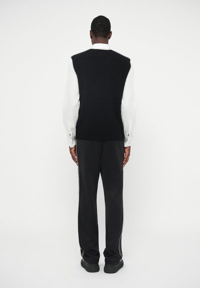 Črn brezrokavnik nad belim dolgim rokavom, v kombinaciji s temnimi hlačami z belimi stranskimi črtami. Gladka tekstura in plasteh zasnova.