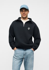 Άνδρας φοράει ανοιχτό μπλε καπέλο, μαύρο μισό-φούτερ με φερμουάρ και λογότυπο στο στήθος, λευκό εσώρουχο και ανοιχτό μπλε τζιν με τα χέρια στις τσέπες.