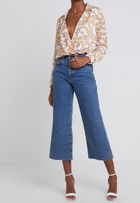 Blouse blanche avec motif de chaînes dorées, décolleté en V profond, associée à des culottes en denim bleu taille haute et des talons blancs à lanières.