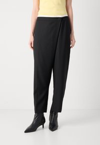 Pantalons noir à jambes larges avec une coupe ajustée, présentant une ceinture contrastante blanche et une texture de tissu lisse et mate. Associés à des bottines pointues.