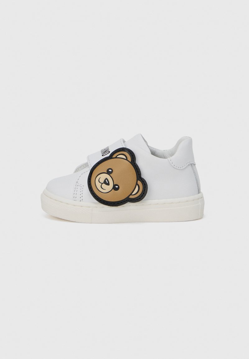 Λευκά αθλητικά παπούτσια με λείο δερμάτινο φινίρισμα, που διαθέτουν στρογγυλό καφέ μπαλώμα αρκούδας στο πλάι και υφασμένο λάστιχο στη σόλα. Κλείσιμο με λουράκι βέλκρο.