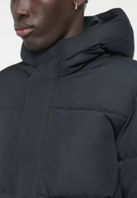 Černá vatovaná bunda s vysokým límcem a kapucí. Má švy na panelech a hladkou matnou texturu. Minimalistický design bez ozdob.