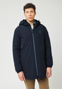 Chaqueta aislante de color navy con capucha, cremallera frontal, puños elásticos y un parche con el logotipo en el pecho. Textura suave y diseño de longitud media.