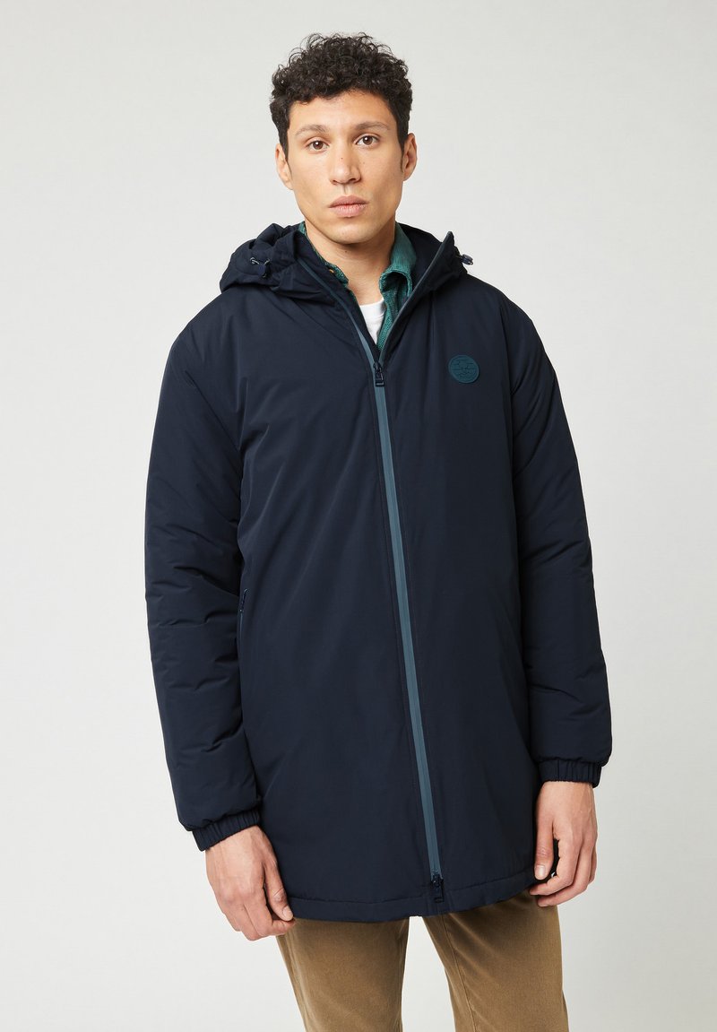 Chaqueta aislante de color navy con capucha, cremallera frontal, puños elásticos y un parche con el logotipo en el pecho. Textura suave y diseño de longitud media.