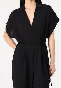 Combinaison portefeuille noire à manches courtes, col en V et ceinture tressée. Réalisée en tissu léger à la texture lisse.