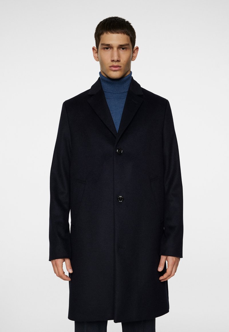 Junger Mann trägt einen marineblauen einreihigen Mantel über einem blauen Rollkragenpullover und steht vor einem schlichten weißen Hintergrund.
