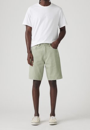 Mand iført plain hvid t-shirt, lysegrønne knælange shorts og beige lærredssko stående mod grå baggrund.