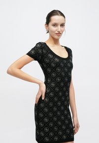 Μαύρο εφαρμοστό φόρεμα με κοντά μανίκια, ντεκολτέ σε σχήμα σκουπ και διακοσμητικό σχέδιο σε σχήμα διαμαντιού με κυκλικά λογότυπα σε αντίθετο χρώμα.