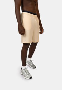 Pantaloni sportivi beige con righe verticali, vita elastica e un piccolo logo di lato. Indossati con scarpe da corsa argentate e calze bianche.