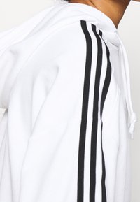 Vit hoodie i en texturerad tygtyp. Har tre svarta ränder längs axeln. Inkluderar en dragsko för justering.