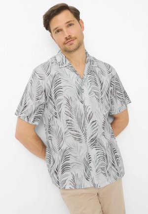 Mand iført lysegrå kortærmet skjorte med mørkt palmebladsmønster og beige bukser, stående mod en hvid baggrund.
