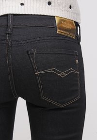 Jeans de mezclilla negra con un corte ajustado, parche de logo dorado en la cinturilla y costuras contrastantes en marrón en los bolsillos y costuras.