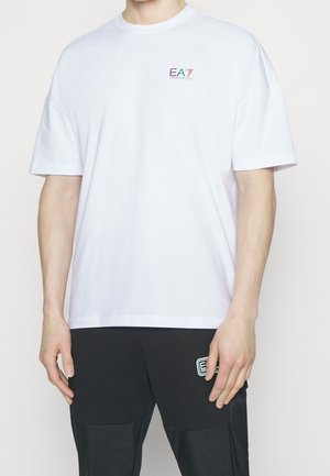 Uomo che indossa una maglietta bianca a maniche corte con un piccolo logo EA7 colorato sul petto e pantaloni neri con un emblema EA7 bianco sulla coscia.