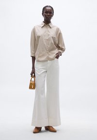 Cămașă bej cu dungi, cu nasturi și mâneci trei sferturi, combinată cu pantaloni albi largi și mule cu toc maro deschis, ținând o geantă mică maro.