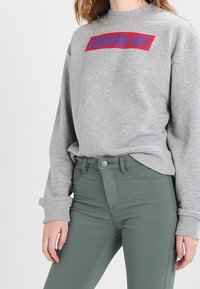 Sudadera gris con texto "SMOKING HOT" en rojo y azul; presenta puños acanalados, cuello redondo y un corte casual, combinada con unos jeans de talle alto verdes.