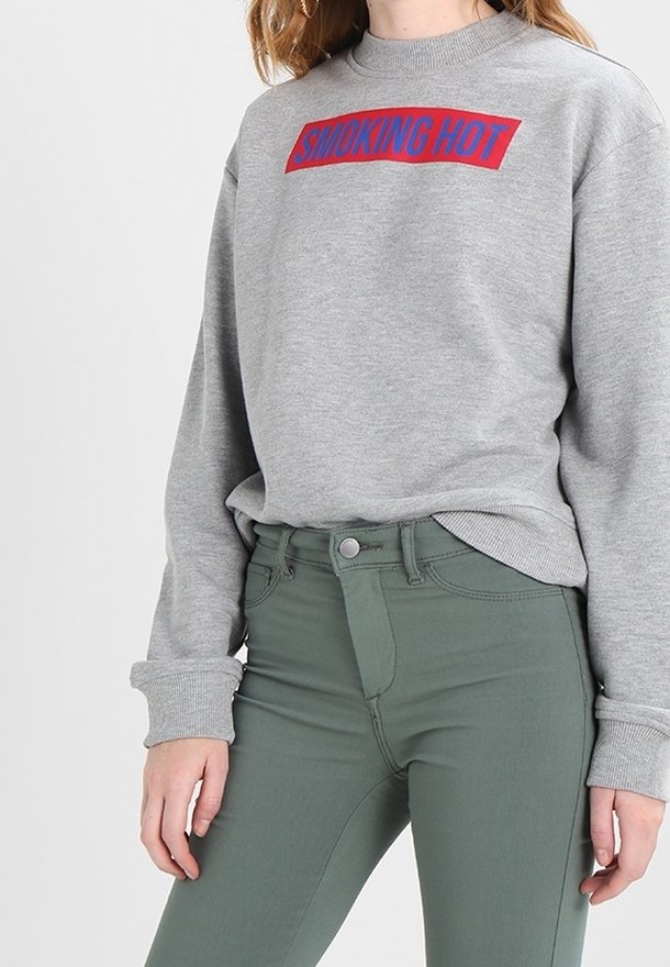 Sudadera gris con texto "SMOKING HOT" en rojo y azul; presenta puños acanalados, cuello redondo y un corte casual, combinada con unos jeans de talle alto verdes.