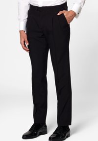Pantalones de vestir negros con un corte ajustado, que presentan pliegues frontales y bolsillos laterales, confeccionados en una tela suave. Combinados con zapatos negros brillantes.