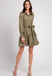 Oliwkowa sukienka z długim rękawem, z kołnierzykiem zapinanym na guziki, z paskiem w talii i warstwowym dołem z falbanami. Wykonana z gładkiego materiału o miękkiej fakturze.